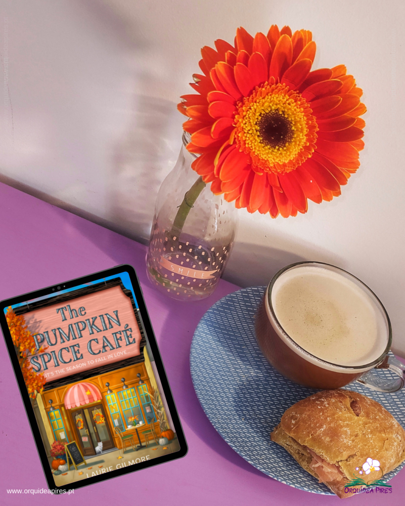 The Pumpkin Spice Café, de Laurie Gilmore, num tablet sobre uma mesa, junto a uma chávena de café, uma sanduíche e uma gerbera vermelha.